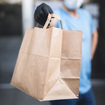 Delivery guy with protective mask and gloves holding box / bag with groceries in front of a building.